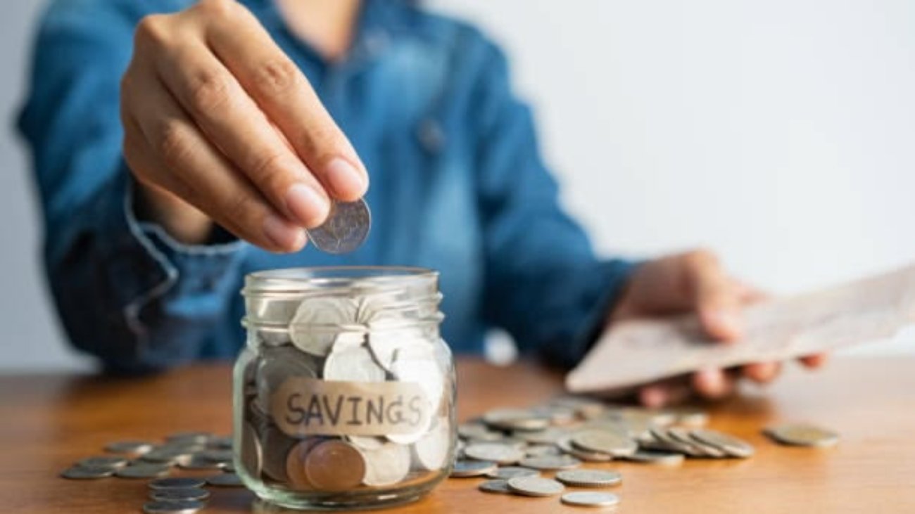 The woman hand is putting a coin in a glass bottle and a pile of coins on a brown wooden table,Investment business, retirement, finance and saving money for future concept.