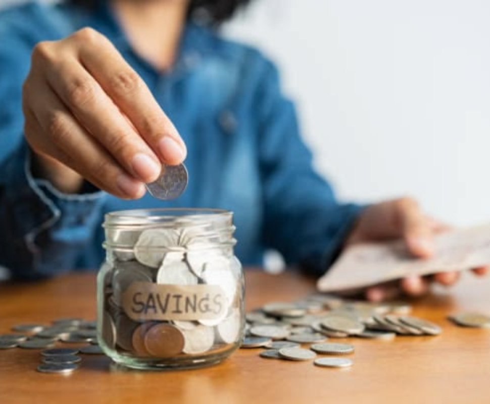 The woman hand is putting a coin in a glass bottle and a pile of coins on a brown wooden table,Investment business, retirement, finance and saving money for future concept.